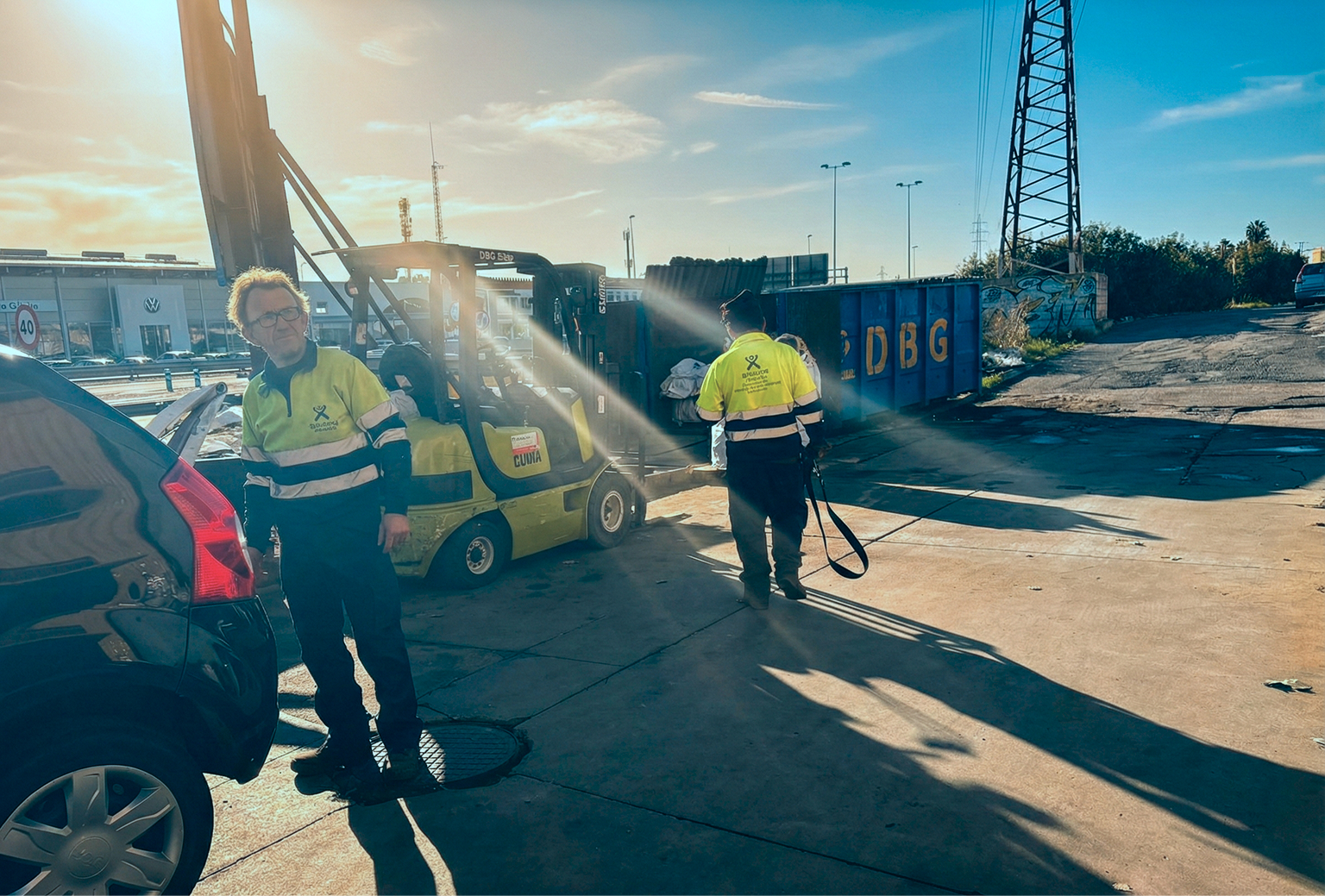 Two workers in yellow and black uniforms load scrap materials using a forklift near a blue container labeled DBG under clear blue sky.