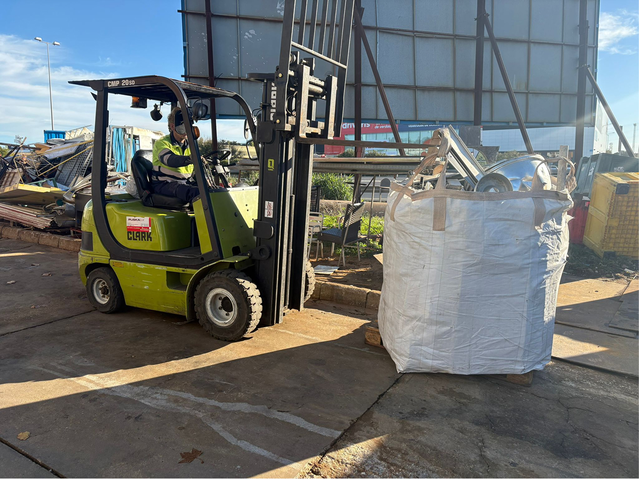 Forklift operated by a worker in safety gear lifting a large white bag filled with metal scrap in an outdoor industrial area.