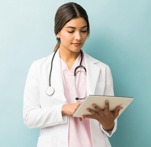Female doctor in white coat and pink scrubs using a tablet with a stethoscope around her neck.