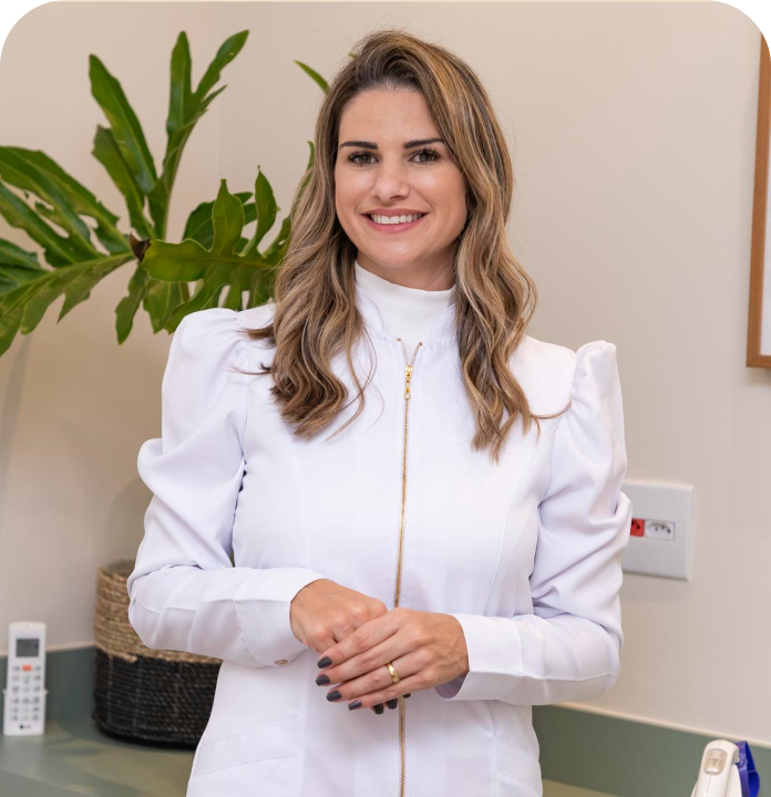 Smiling woman with long wavy blonde hair wearing a white zip-up medical or dental jacket standing in a room with a plant in the background.