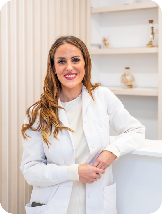 Smiling woman in a white medical coat leaning on a white counter with shelves in the background.