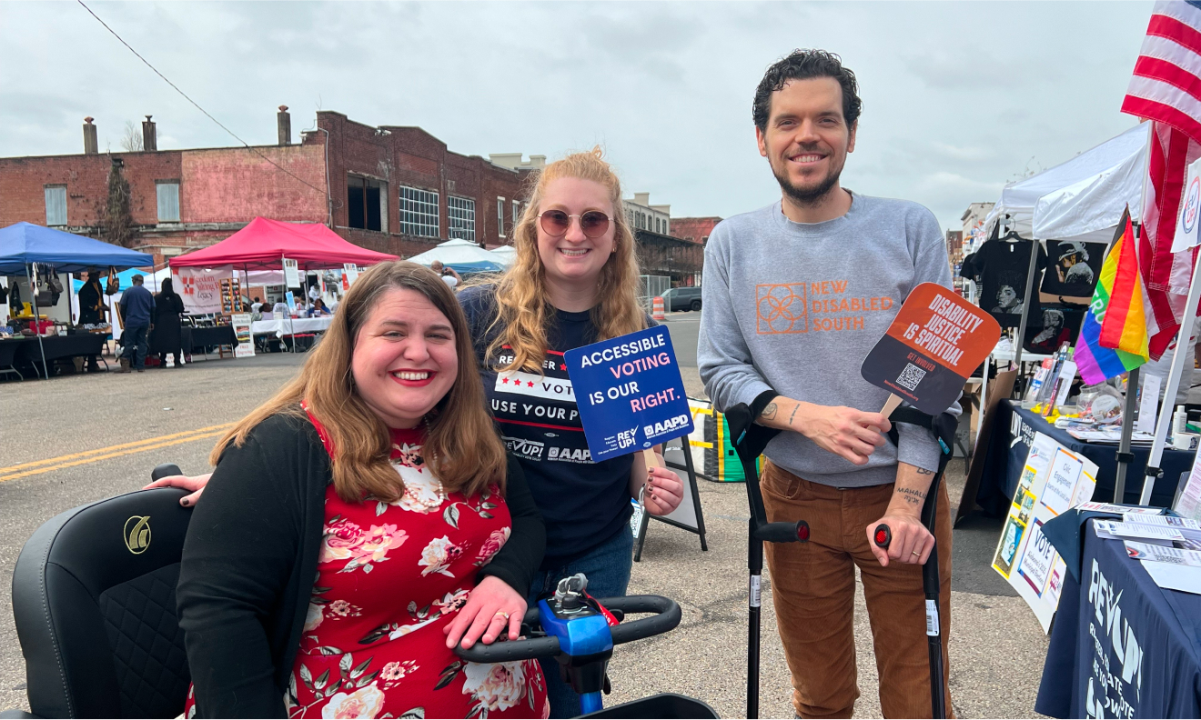 Three people pose together on a street with vendor tents behind them. The person on the left is seated on a mobility scooter, the person in the middle holds a sign that reads “Accessible voting is our right,” and the person on the right stands using crutches and holds a sign that reads “Disability justice is spiritual.”