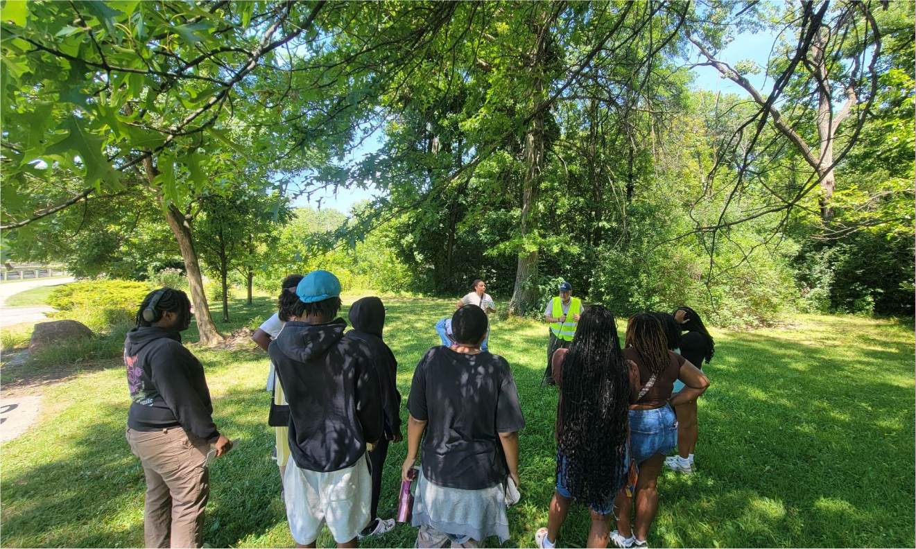 A group of people stand together on a grassy area in a wooded park, listening to two speakers standing in front of them. Large trees with green leaves hang overhead, and sunlight filters through the branches.