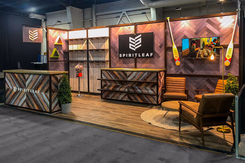 10x20 linear modular trade show booth for a Cannabis retailer. Wooden floors fill the booth space with textured herringbone wooden wall panels. Multi-coloured wooden counters in triangular slat patterns are displayed in the middle and to the left. The back