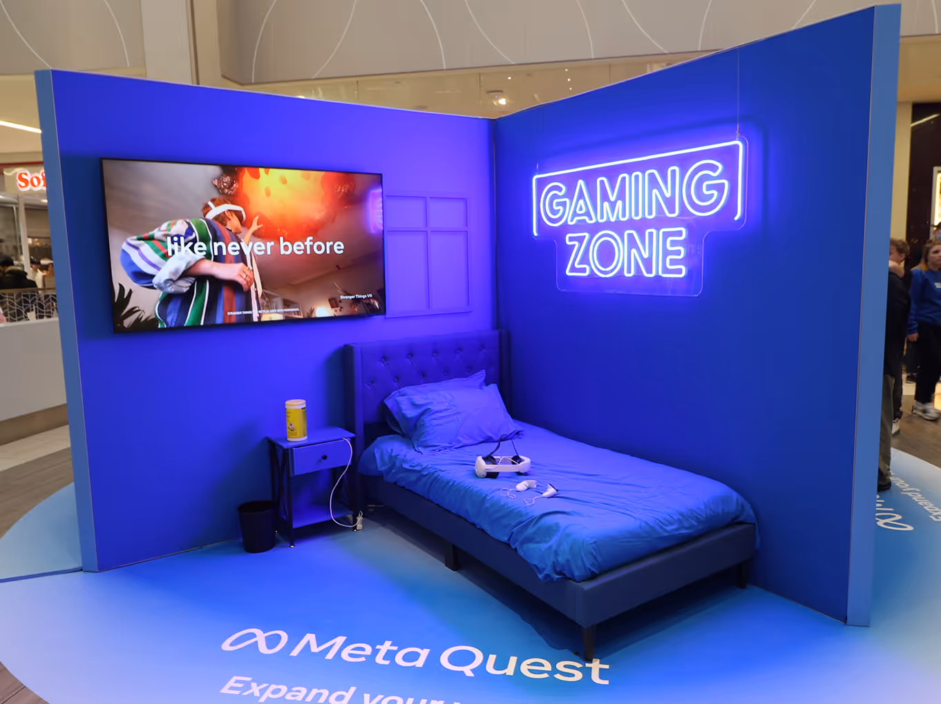 open-concept VR zone display showing a bed, wall-mounted tv, and "gaming zone" LED sign with a branded blue floor decal