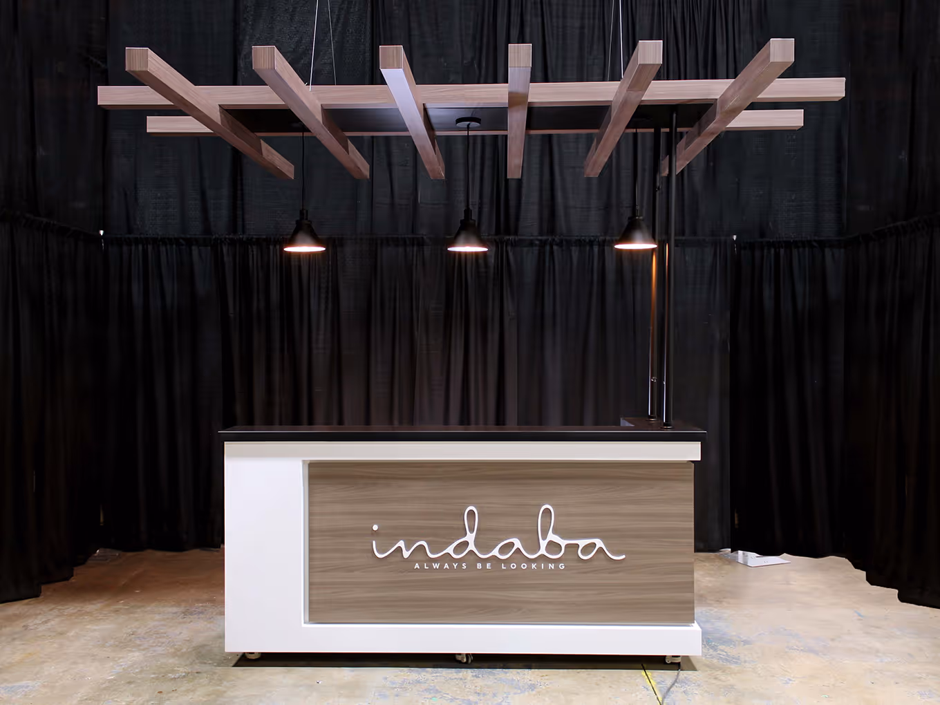 Completed Indaba reception counter with wood-grain front panel, illuminated logo, and overhead canopy with integrated pendant lighting.