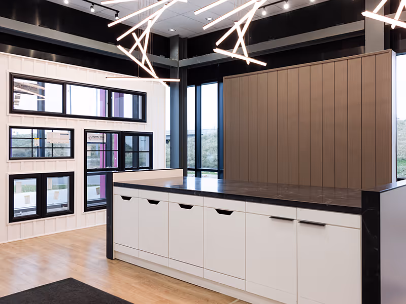 Showroom island counter with black Meganite countertop, integrated storage drawers, and a full wall display of Ply Gem window samples.