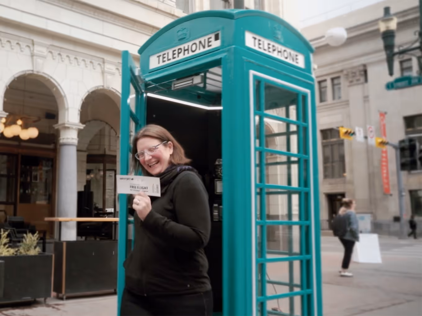 Winner stepping out of the teal WestJet phone booth on Stephen Avenue holding a free flight boarding pass to London.