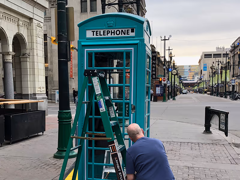 Technician installing and wiring the teal English phone booth on Stephen Avenue for WestJet’s Calgary-to-London campaign.
