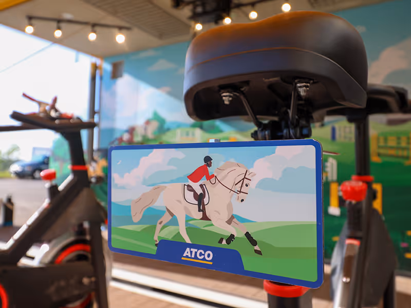 Close-up of an ATCO-branded race-themed illustration mounted on a stationary bike inside the Pedal Prix interactive cycling game container.