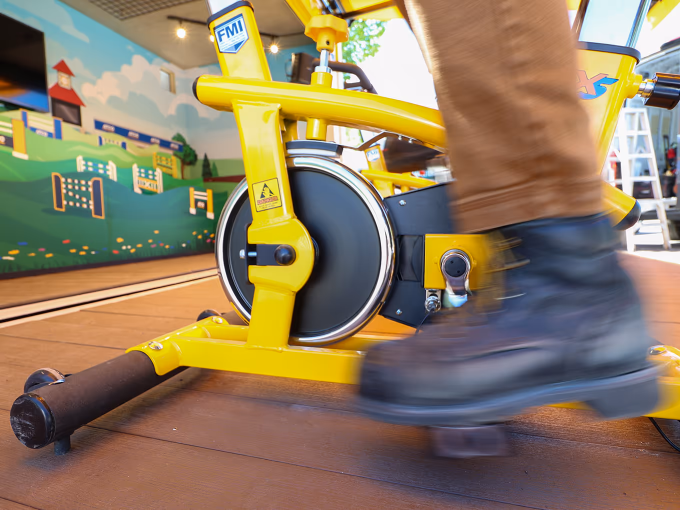 Close shot of stationary bike pedaling, featuring an Interior wall mural with 3D horse jump graphics and mounted monitor inside custom container display.