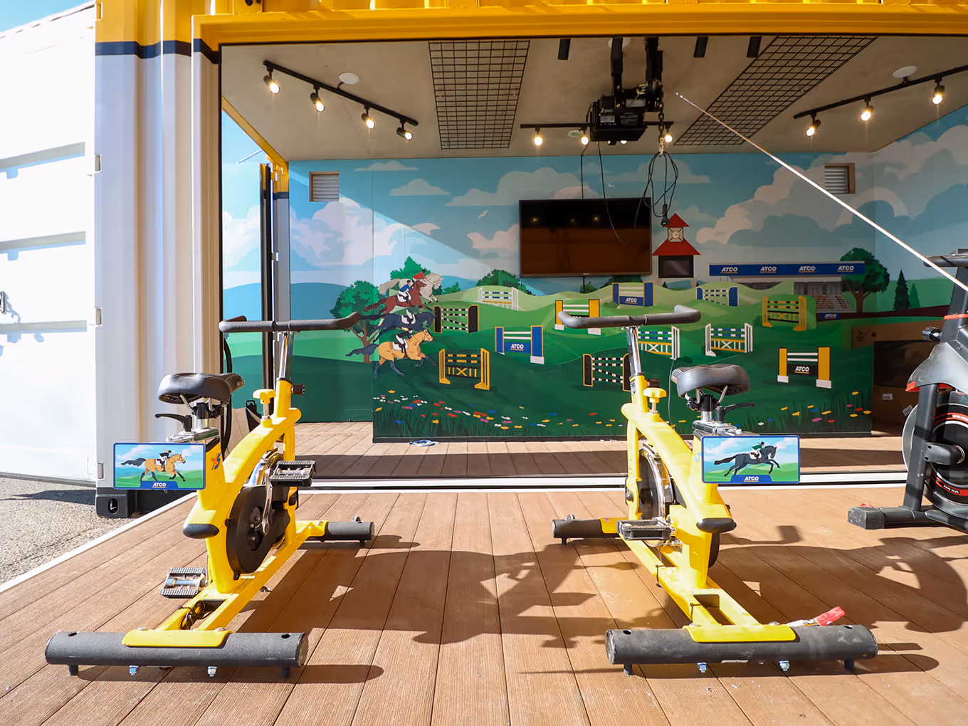 Inside view of ATCO Pedal Prix activation showing interactive horse-race game wall graphics and stationary bikes in custom shipping container.