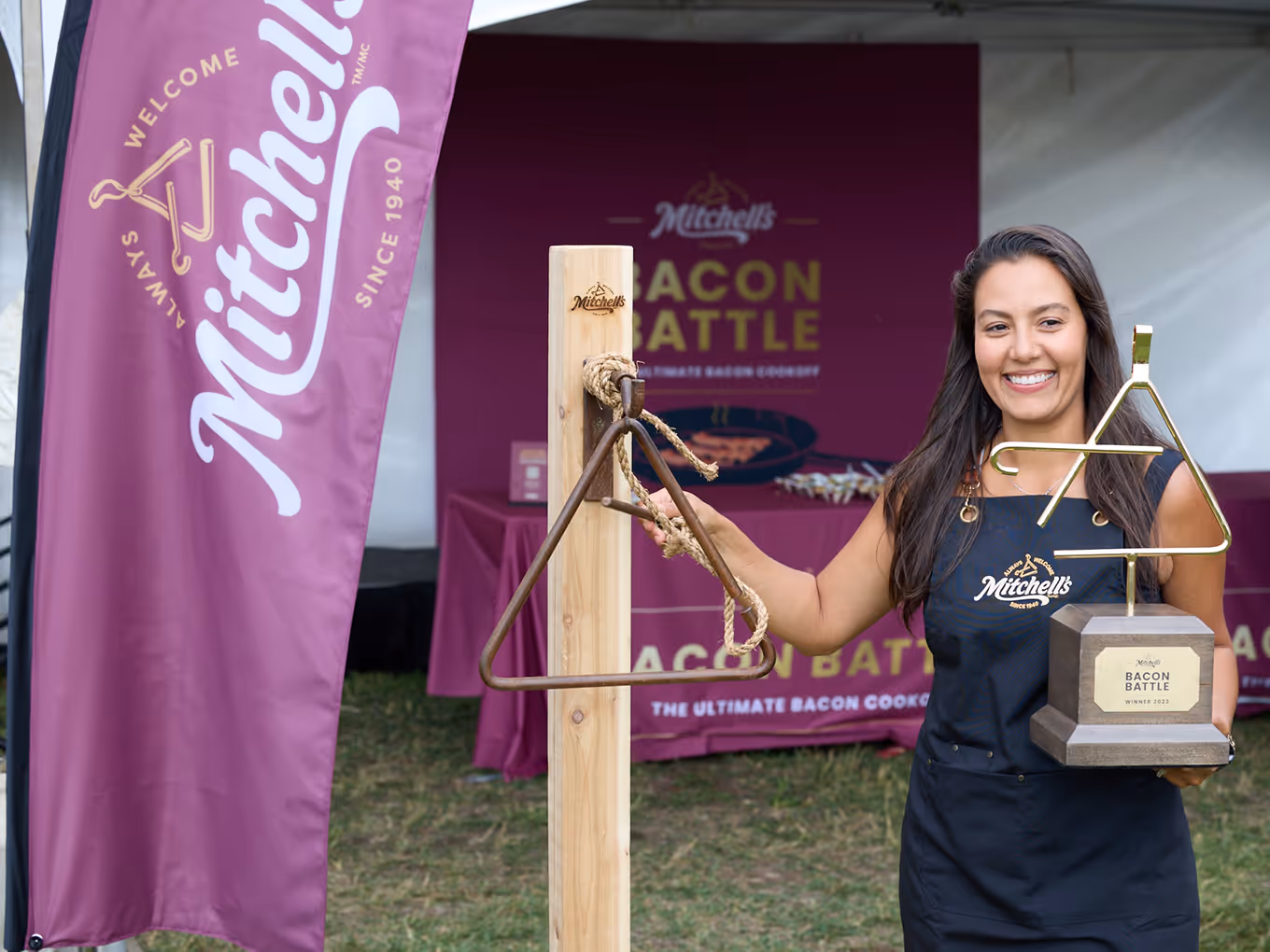 Winner holding the Mitchell’s Bacon Battle custom gold-plated trophy next to branded event displays and cedar dinner bell.