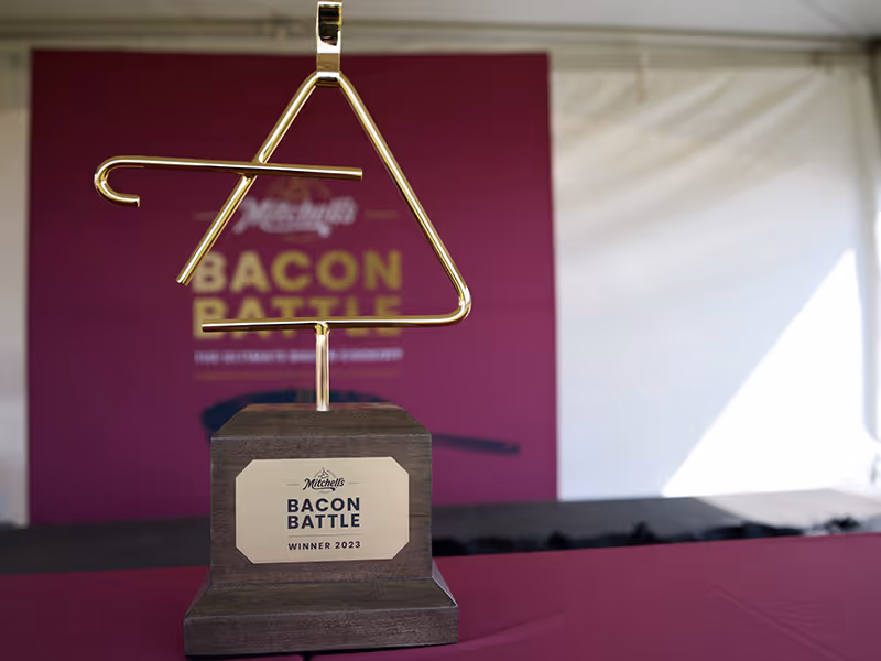 Custom gold-plated Mitchell’s Bacon Battle trophy on a walnut-stained maple base displayed in front of the portable fabric branded event backdrop.