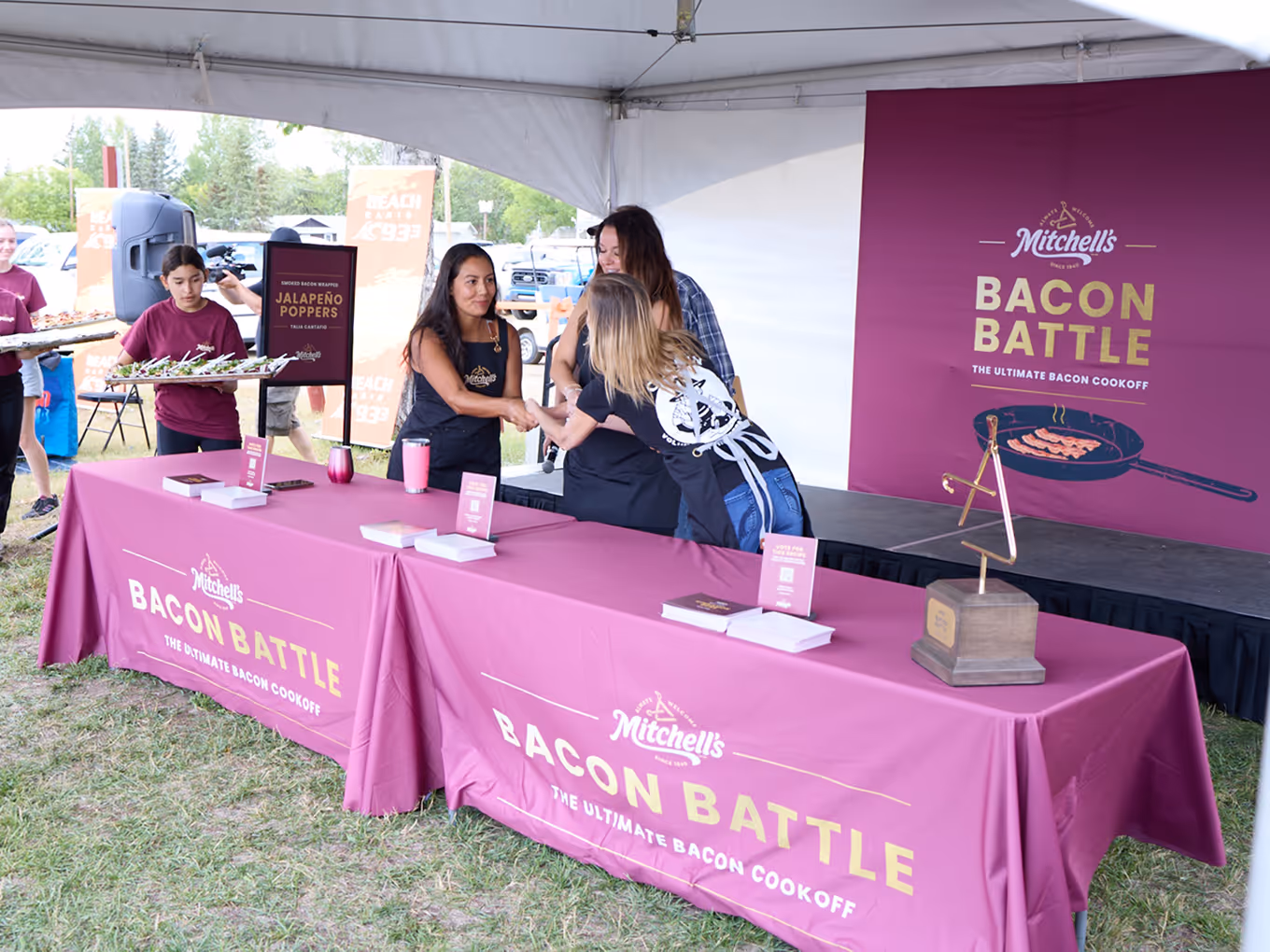 Mitchell’s Bacon Battle event setup featuring branded flags, tablecloths, and competitors preparing dishes for the live cook-off.