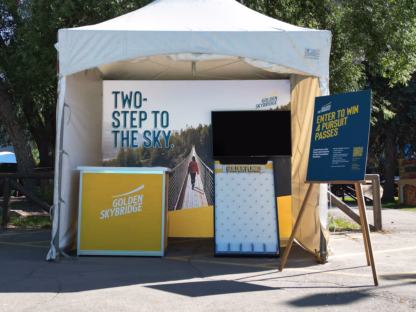 Outdoor Golden Skybridge booth with branded backdrop, counter, Plinko game, and contest sign under a white tent.