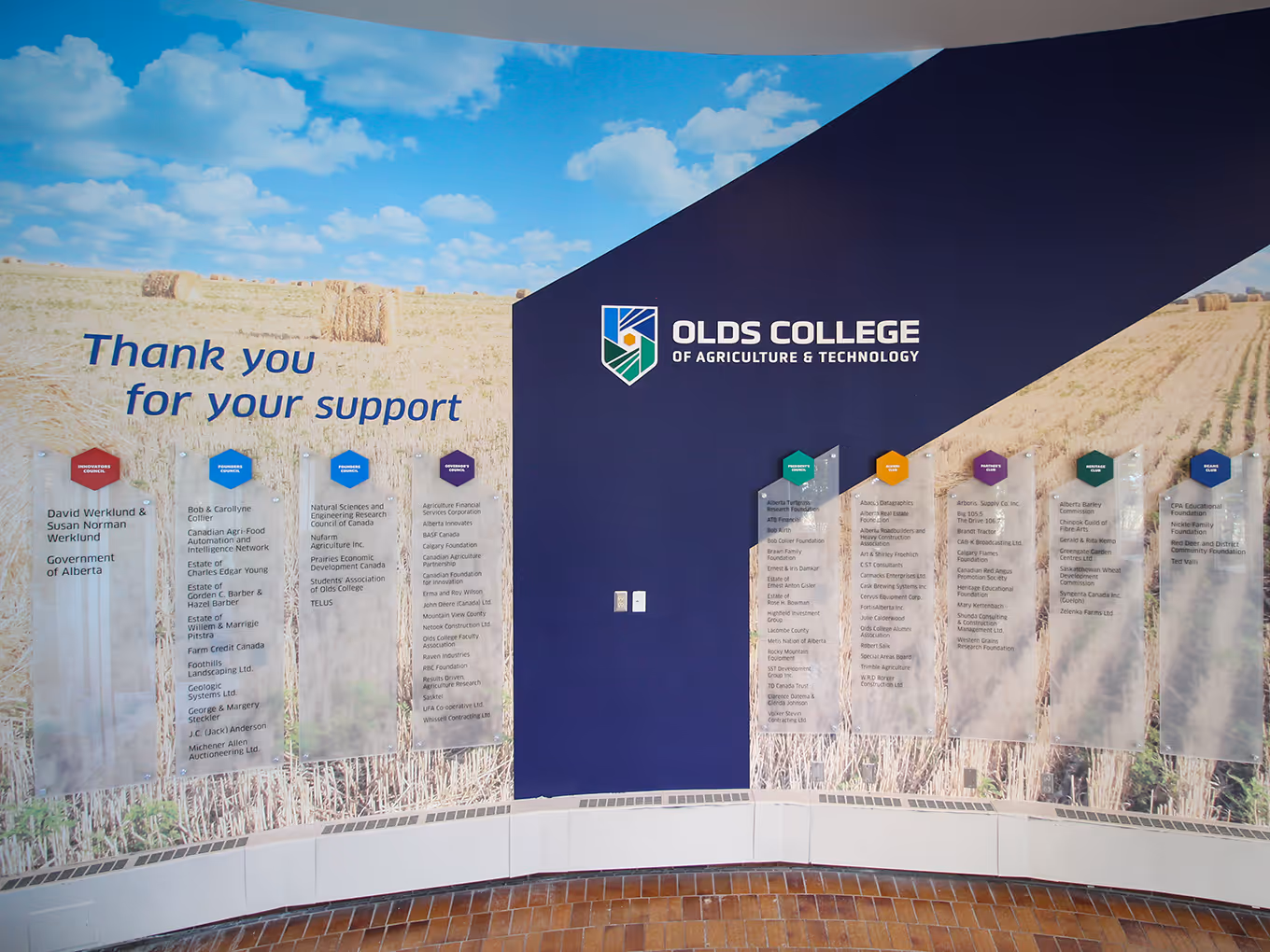 Wide view of the Olds College donor wall featuring full-height vinyl graphics, branded lettering, and frosted acrylic donor panels on a curved wall.