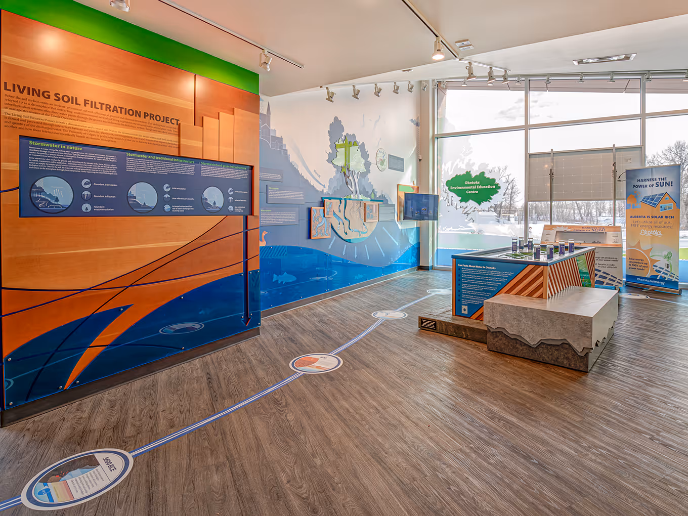 Wide interior view of the Okotoks Interpretive Centre highlighting the Living Soil Filtration Project exhibit, interactive displays, and guided floor pathway.