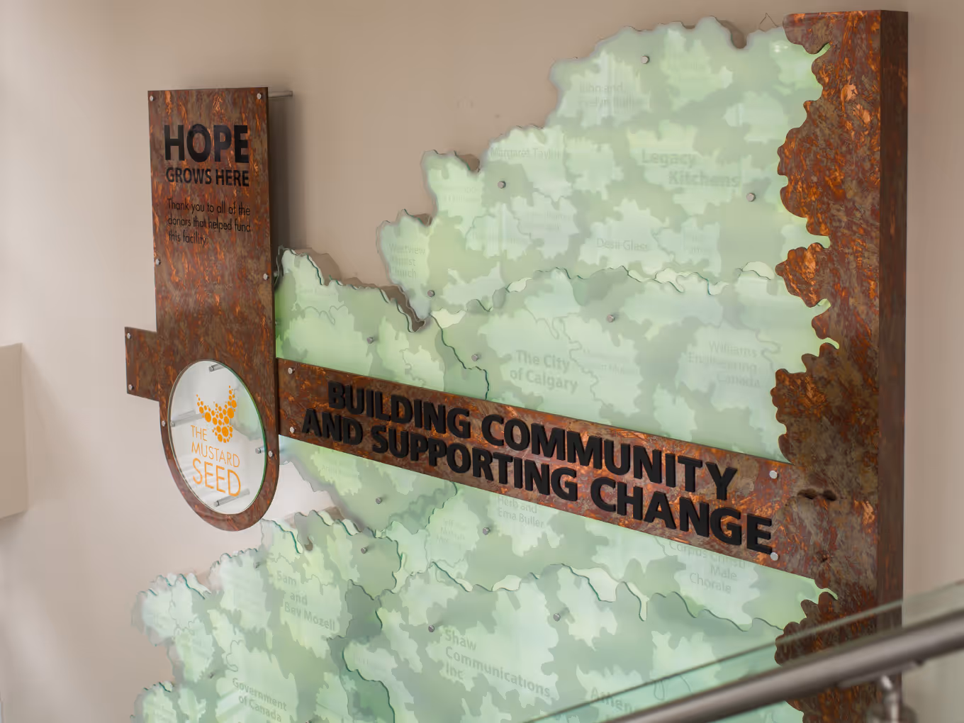 Close-up of The Mustard Seed donor wall showing frosted acrylic layers with etched donor names and copper framing.