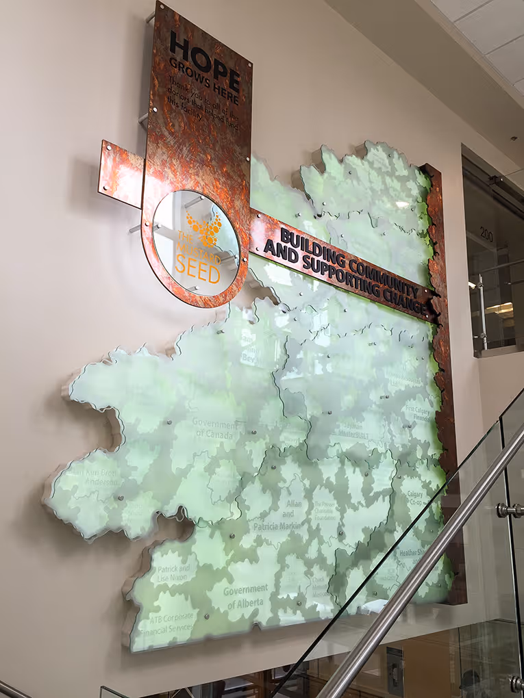 Side view of The Mustard Seed donor recognition wall integrated into a stairwell, featuring layered frosted acrylic panels with etched donor names and copper framing.