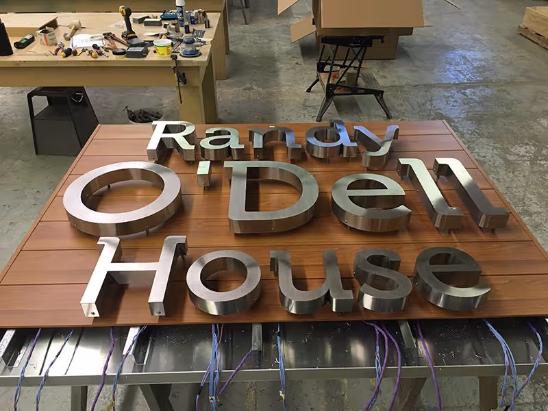 Halo-lit exterior signage for Randy O’Dell House, showing dimensional chrome lettering mounted to a wood-clad wall panel during fabrication.