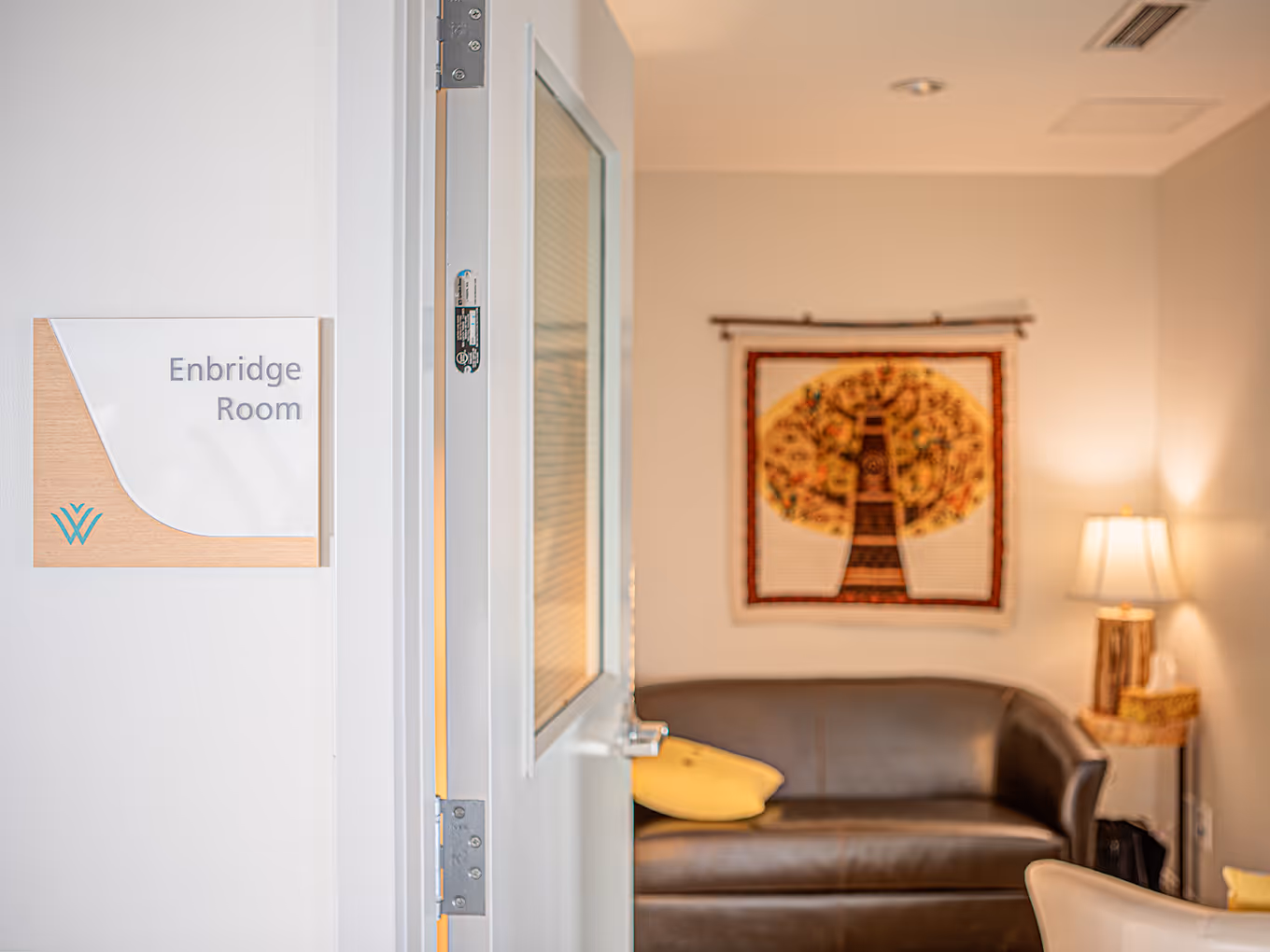 Enbridge Room sign with frosted acrylic and wood backing mounted beside a doorway leading into a lounge.