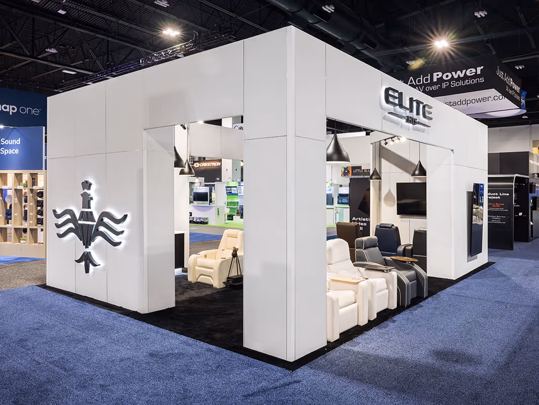 White modular trade show booth with illuminated Elite branding, open entryways, product displays, and lounge seating on a dark carpeted floor.