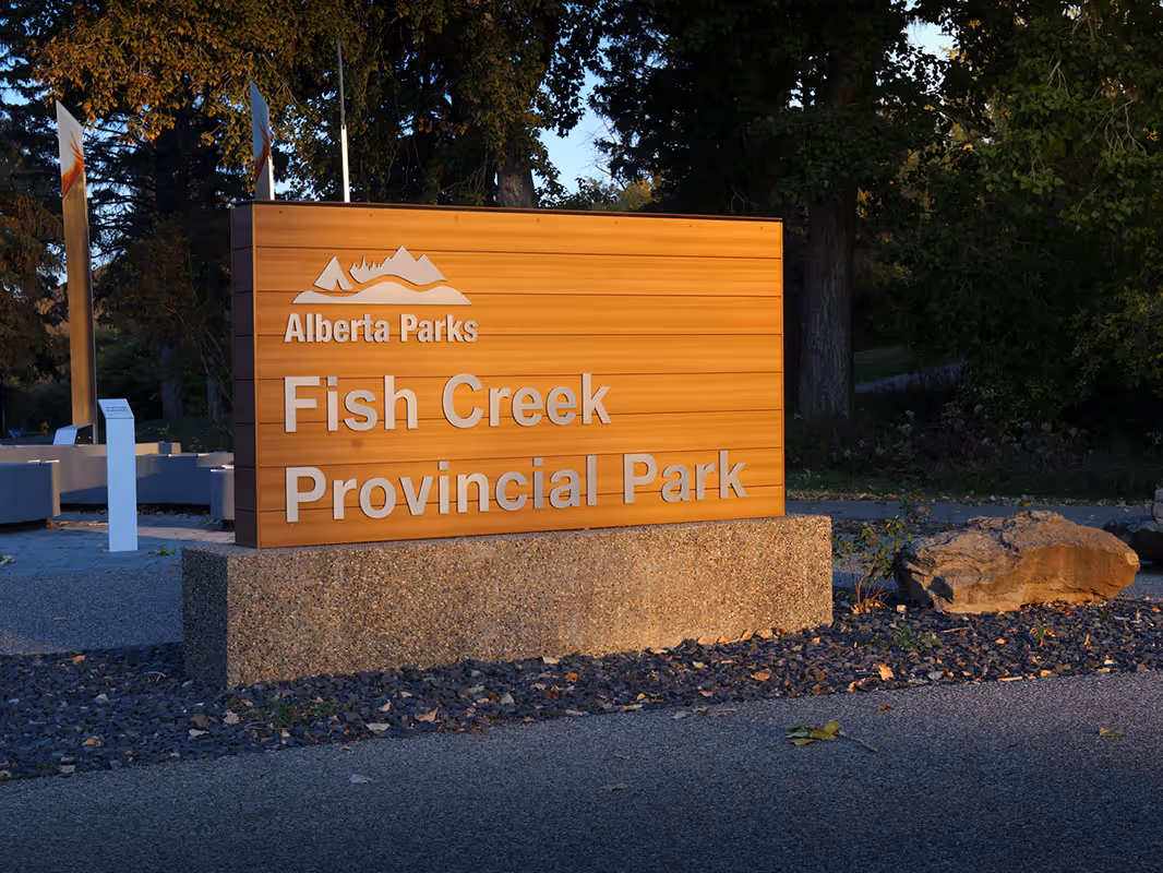 Wood-paneled Alberta Parks sign for Fish Creek Provincial Park, mounted on a concrete base at the park entrance with trees in the background.