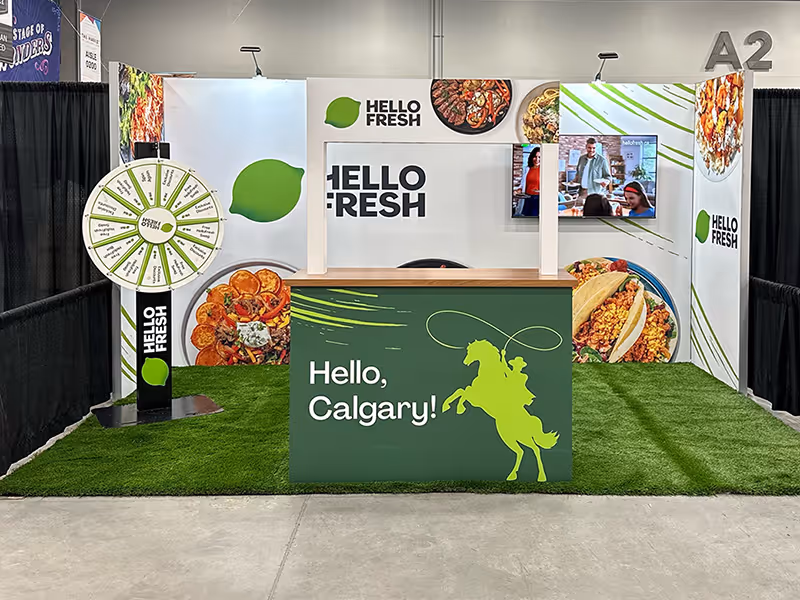 HelloFresh 10x20 Calgary Stampede market booth with custom counter, prize wheel, fabric wall graphics, wall-mounted monitor, and grass carpet display.