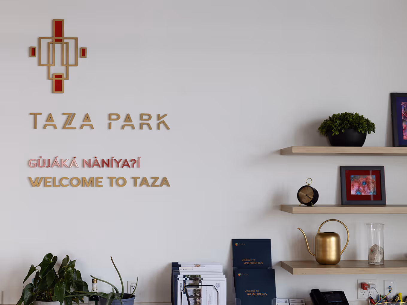 Taza Park branded wall signage with gold logo and "Welcome to Taza" in Tsuut'ina and English, with styled shelving and brochures below.