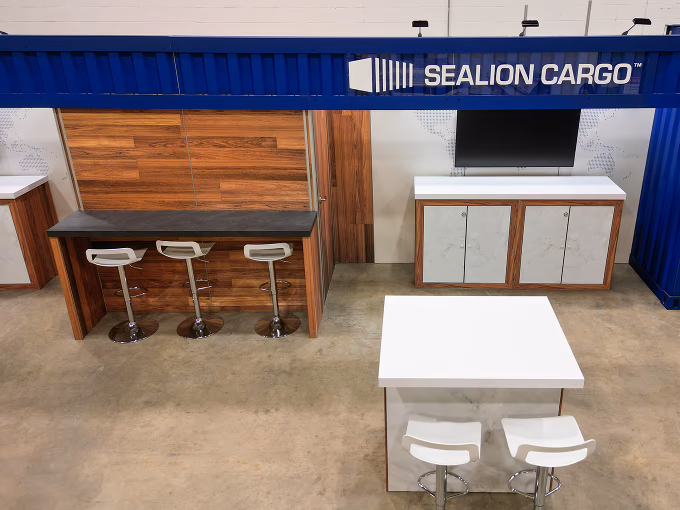 Modern trade show booth with wood finishes, white meeting table, bar seating, and built-in display for product presentations.