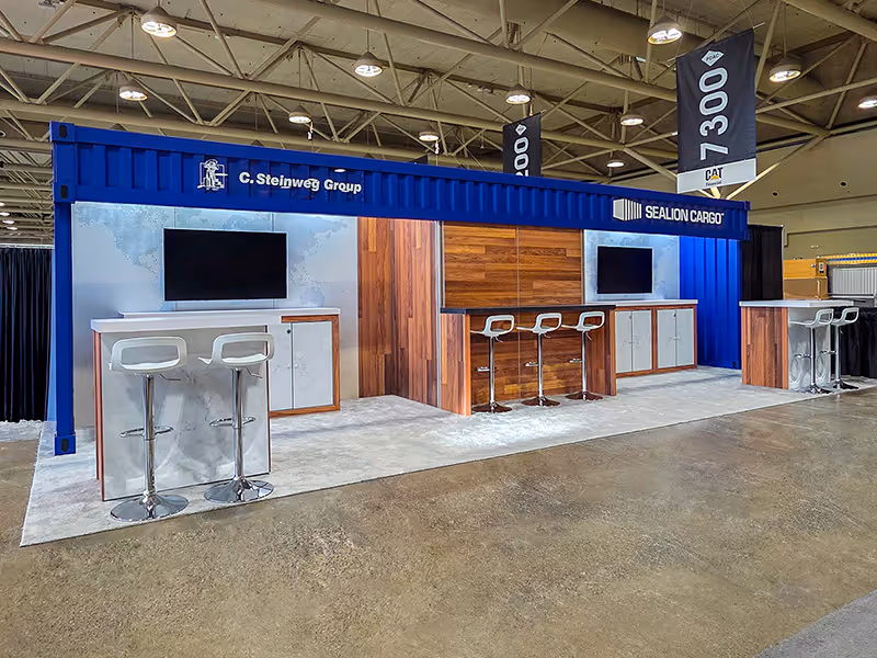Branded 10x30 shipping container exhibit with wood paneling, demo counters, seating, and overhead signage on show floor.