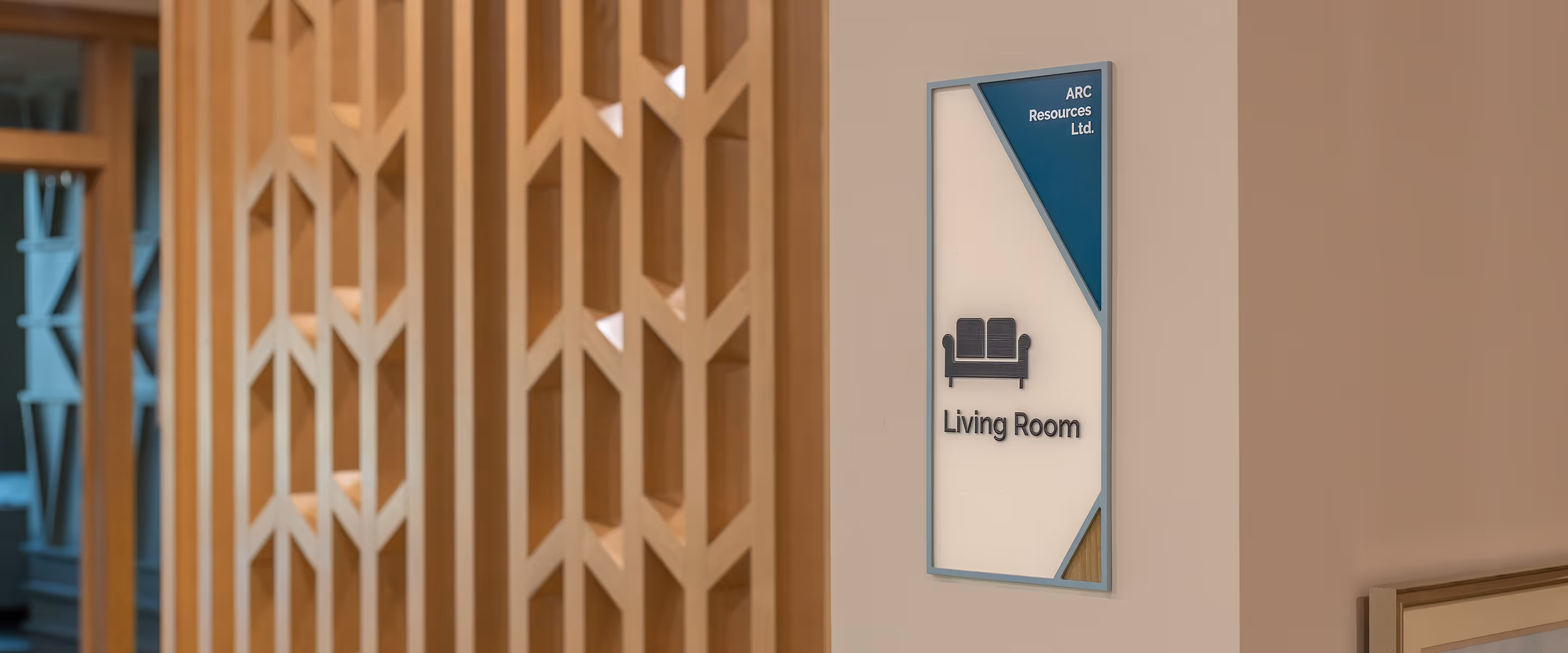 Custom room identification sign for the Living Room at Ronald McDonald House Alberta Calgary House, featuring a painted blue-grey acrylic frame, teal sponsor corner for ARC Resources Ltd., dimensional sofa icon, wood grain graphic panel, and braille, mount
