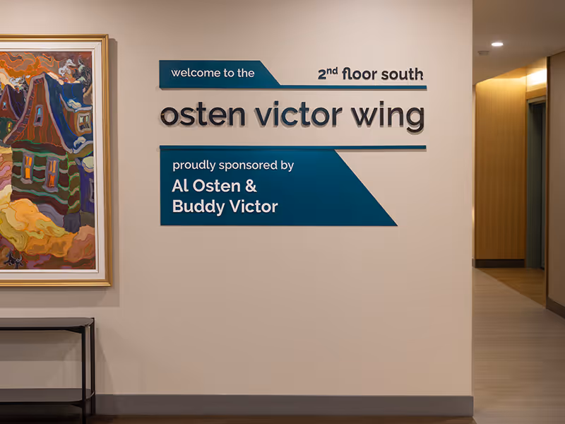 nstalled building wing identification signage at Ronald McDonald House Alberta Calgary House, featuring dimensional black 3D letters spelling "osten victor wing," layered teal painted acrylic panels reading "welcome to the 2nd floor south" and "proudly spo