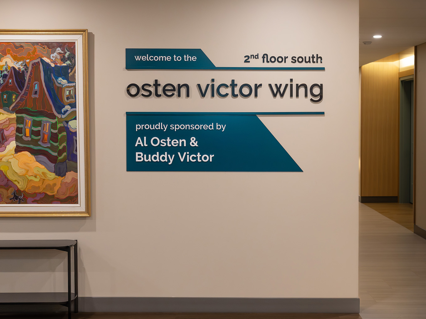 Installed building wing identification signage at Ronald McDonald House Alberta Calgary House, featuring large-scale dimensional black 3D letters spelling "osten victor wing," layered teal painted acrylic panels reading "welcome to the 2nd floor south" and