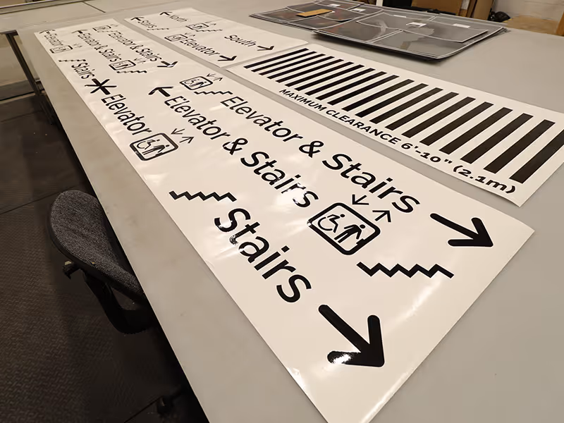 Large-format wayfinding sign graphics laid out on a production table at Exhibit Studio, showing print-ready elevator, stairs, and maximum clearance directional signage with bold black text, accessibility icons, and directional arrows on white acrylic panel