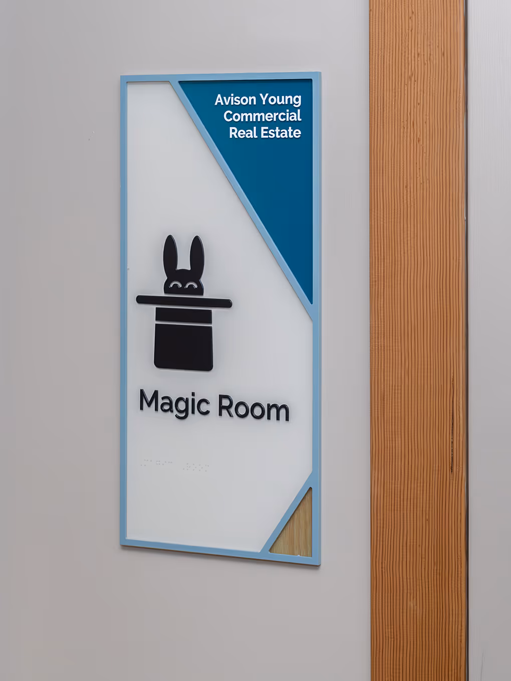 Custom wayfinding room ID sign for the Magic Room at Ronald McDonald House Alberta Calgary House, featuring a painted blue acrylic frame, teal sponsor corner for Avison Young Commercial Real Estate, dimensional rabbit-in-hat icon, braille panel, and wood g
