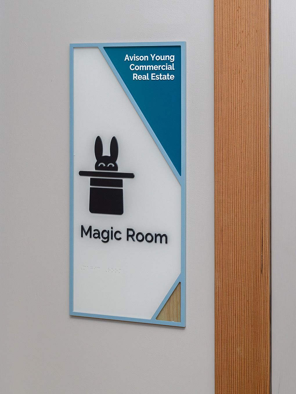 Custom wayfinding room ID sign for the Magic Room at Ronald McDonald House Alberta Calgary House, featuring a painted blue acrylic frame, teal sponsor corner for Avison Young Commercial Real Estate, dimensional rabbit-in-hat icon, braille panel, and wood g