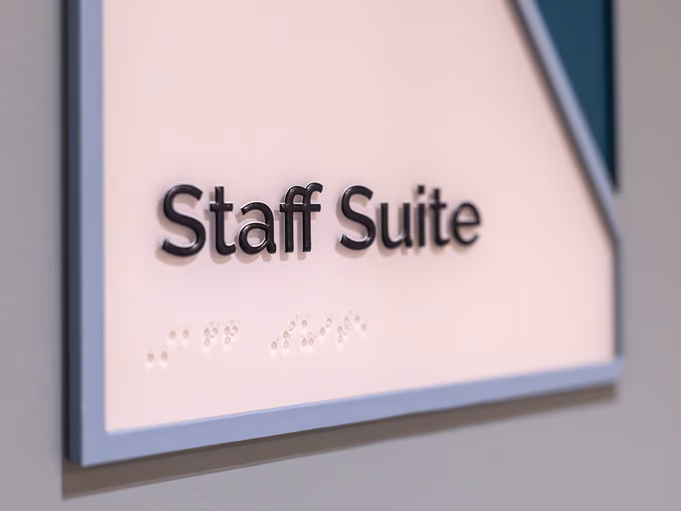 Close-up detail of a custom accessible room identification sign at Ronald McDonald House Alberta Calgary House, showing dimensional black 3D letters spelling "Staff Suite," a braille panel below, painted blue-grey acrylic frame, and teal sponsor corner gra