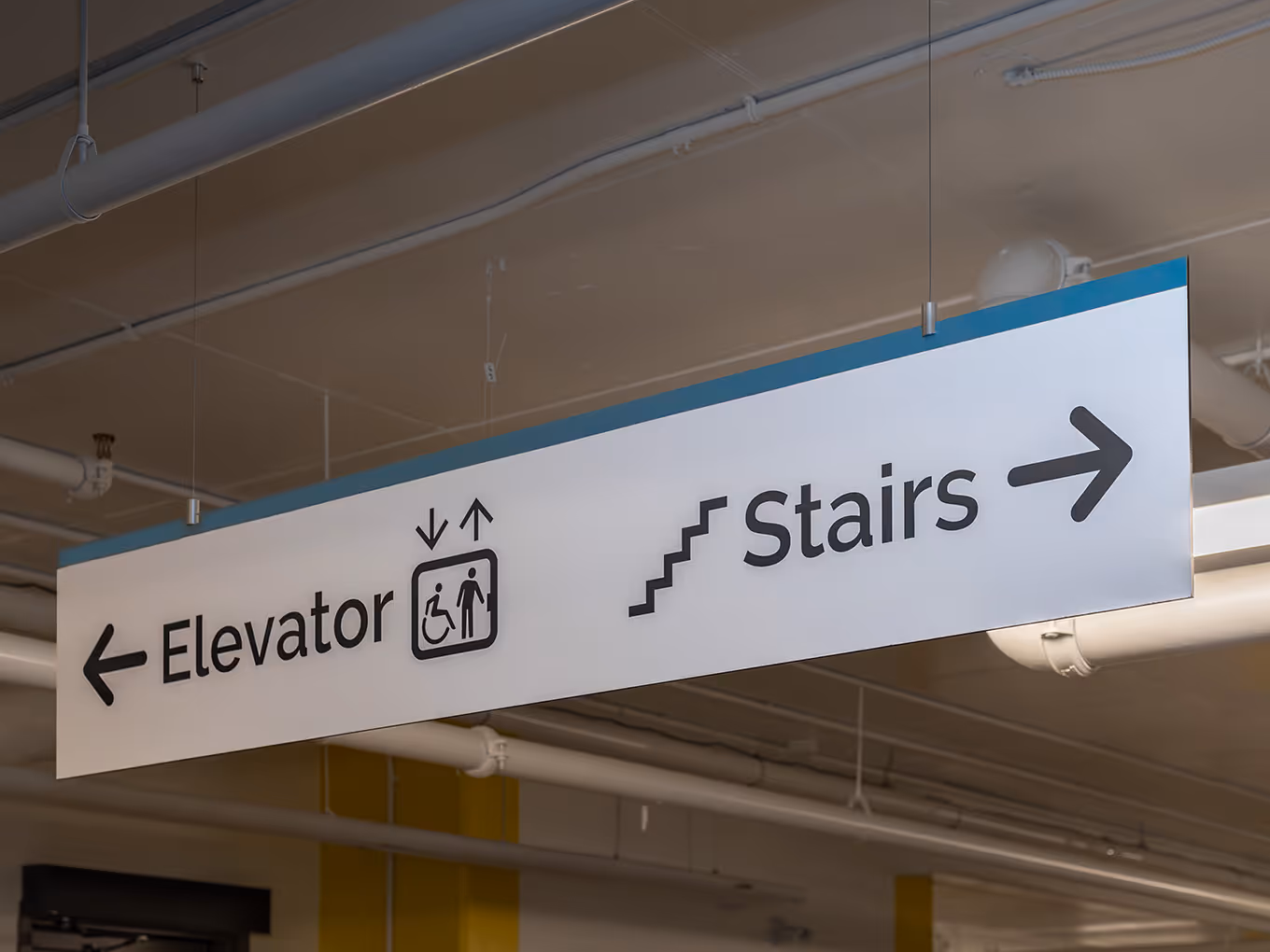 Suspended parkade wayfinding sign at Ronald McDonald House Alberta Calgary House, directing to elevator and stairs with accessibility icons.