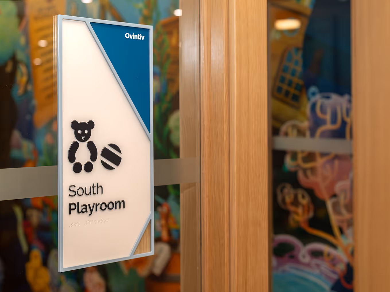 Room ID sign for South Playroom at RMHC Calgary House, featuring dimensional teddy bear icon, braille panel, teal sponsor corner for Ovintiv, and acrylic frame.