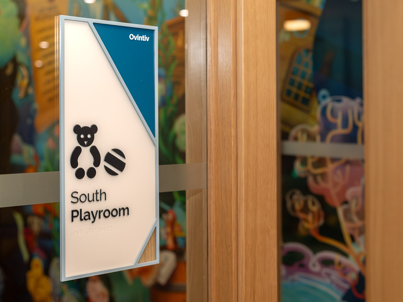 Room ID sign for South Playroom at RMHC Calgary House, featuring dimensional teddy bear icon, braille panel, teal sponsor corner for Ovintiv, and acrylic frame.