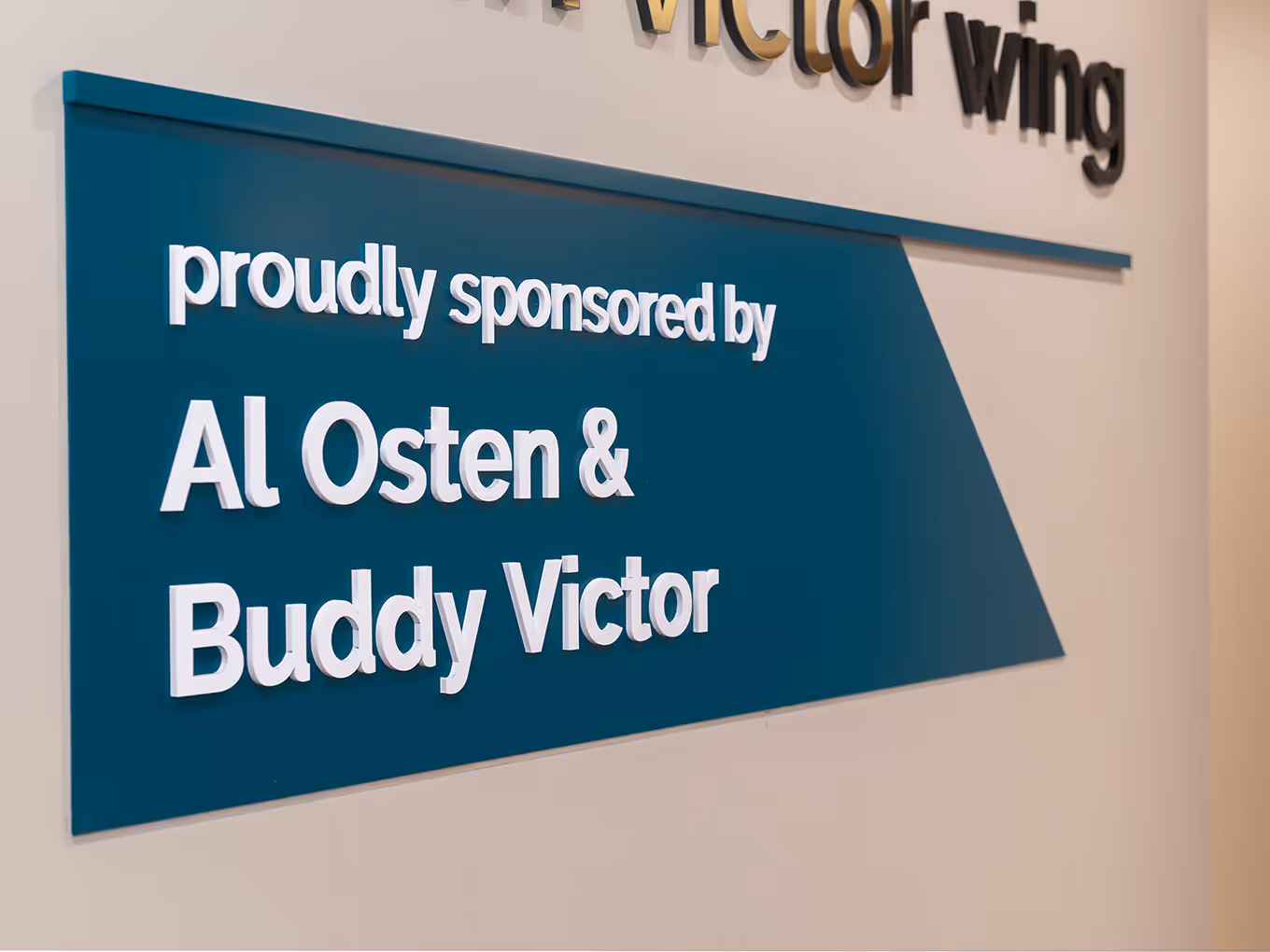 Close-up of dimensional white 3D lettering on a teal painted acrylic sponsor panel reading "Al Osten & Buddy Victor" at Ronald McDonald House Alberta.
