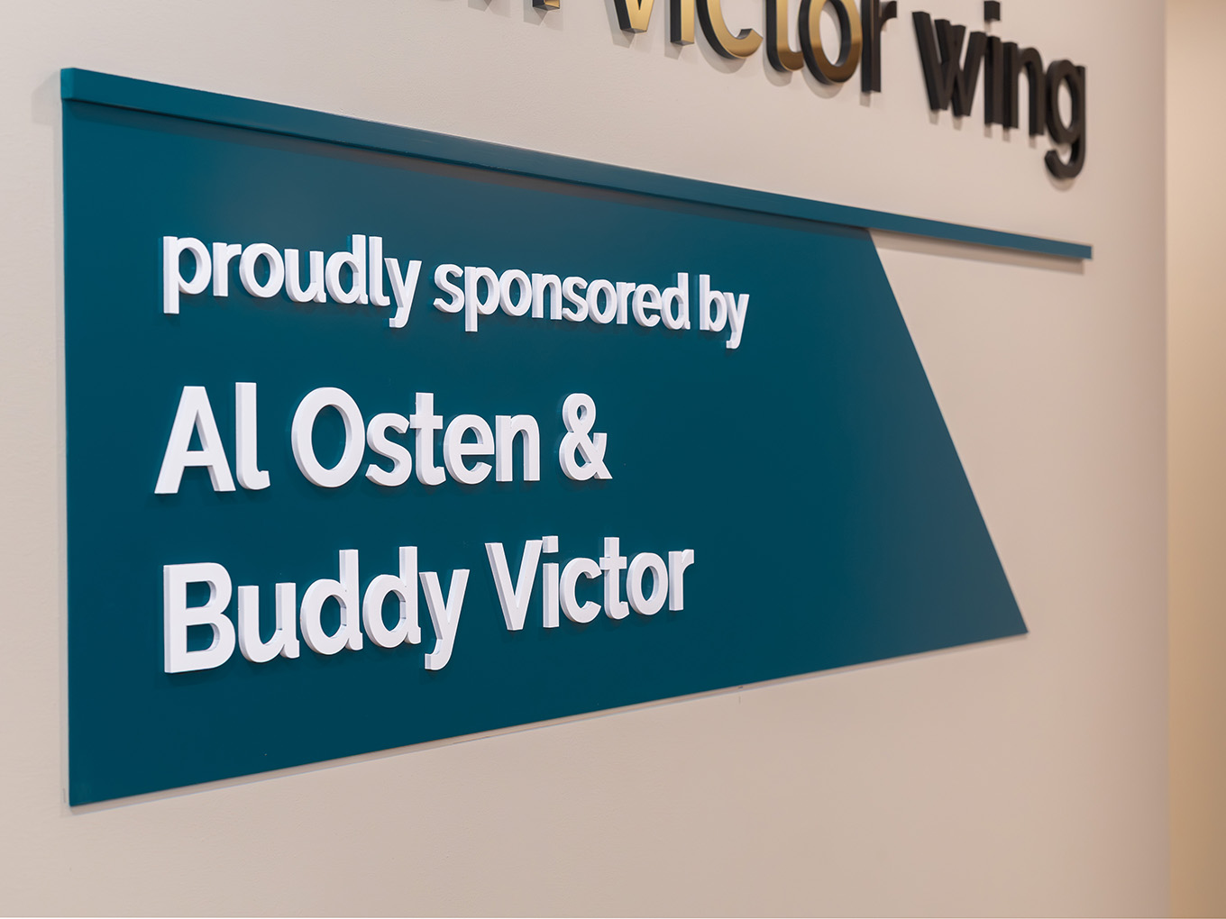 Close-up of dimensional white 3D lettering on a teal painted acrylic sponsor panel reading "Al Osten & Buddy Victor" at Ronald McDonald House Alberta.