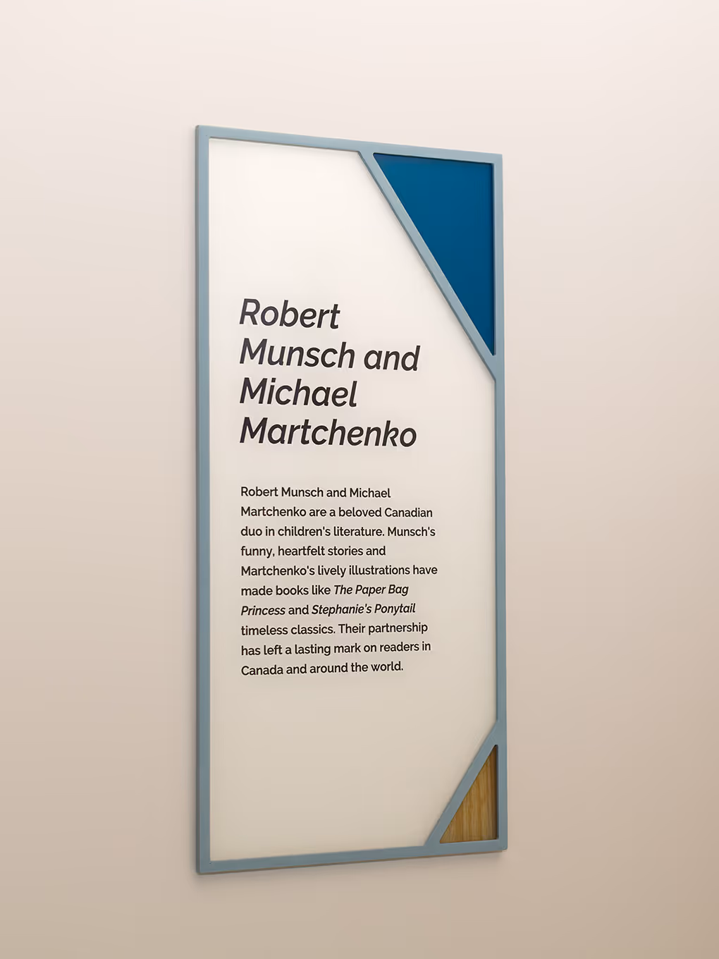 Custom interpretive wall sign at RMHC Calgary House featuring painted acrylic frame, teal and wood grain corner graphics, and text about Robert Munsch and Michael Martchenko.