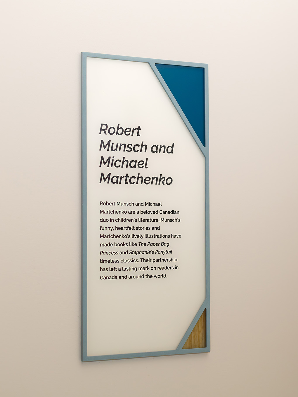 Custom interpretive wall sign at RMHC Calgary House featuring painted acrylic frame, teal and wood grain corner graphics, and text about Robert Munsch and Michael Martchenko.