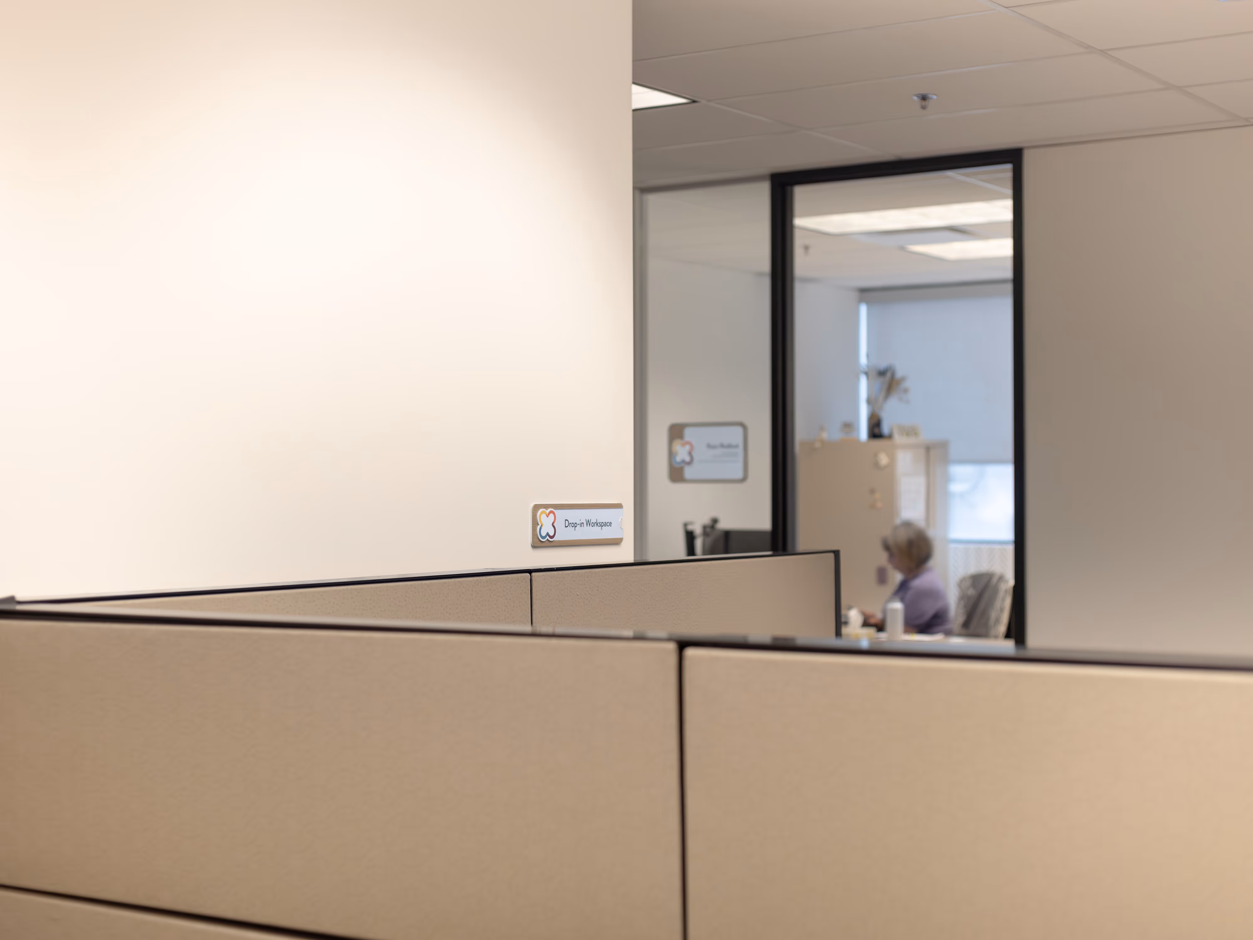 A small Drop-in Workspace plaque mounted on a low office partition, with cubicles in the foreground and a staff member working at a desk in the background.