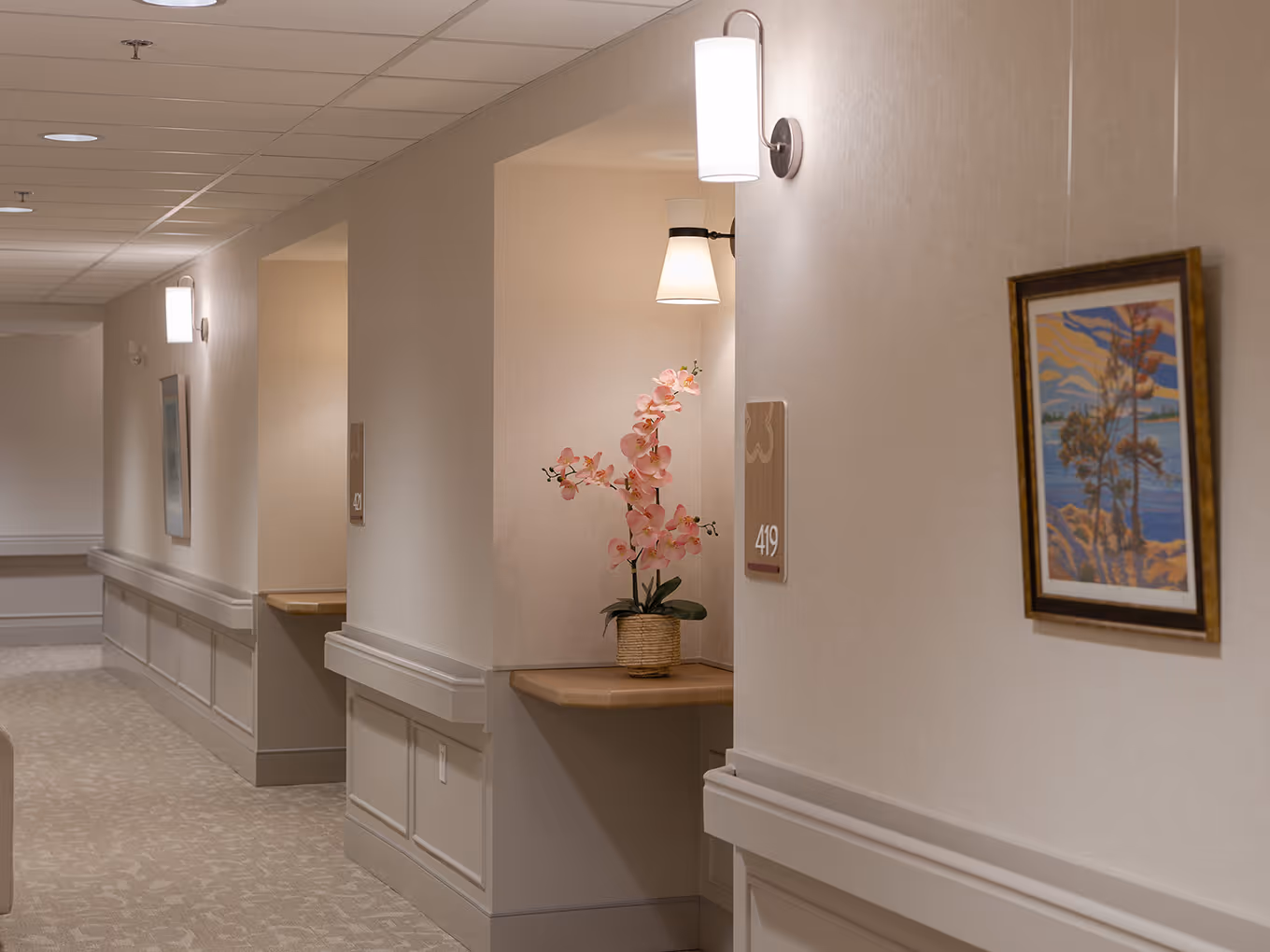 Warm residential corridor at United Active Living Garrison Green featuring custom suite signage, soft sconce lighting, and locally inspired artwork — a calm, easy-to-navigate environment.