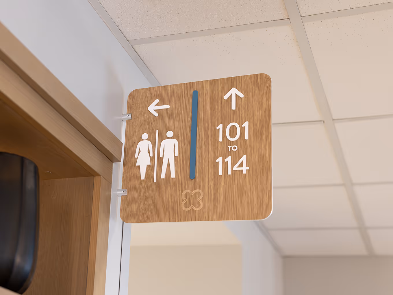 Wood-look wall-mounted directional sign at United Active Living Garrison Green pointing toward suites 101–114 and the washrooms, featuring dimensional arrows, washroom icons, a blue accent bar, and the engraved United floral logo.