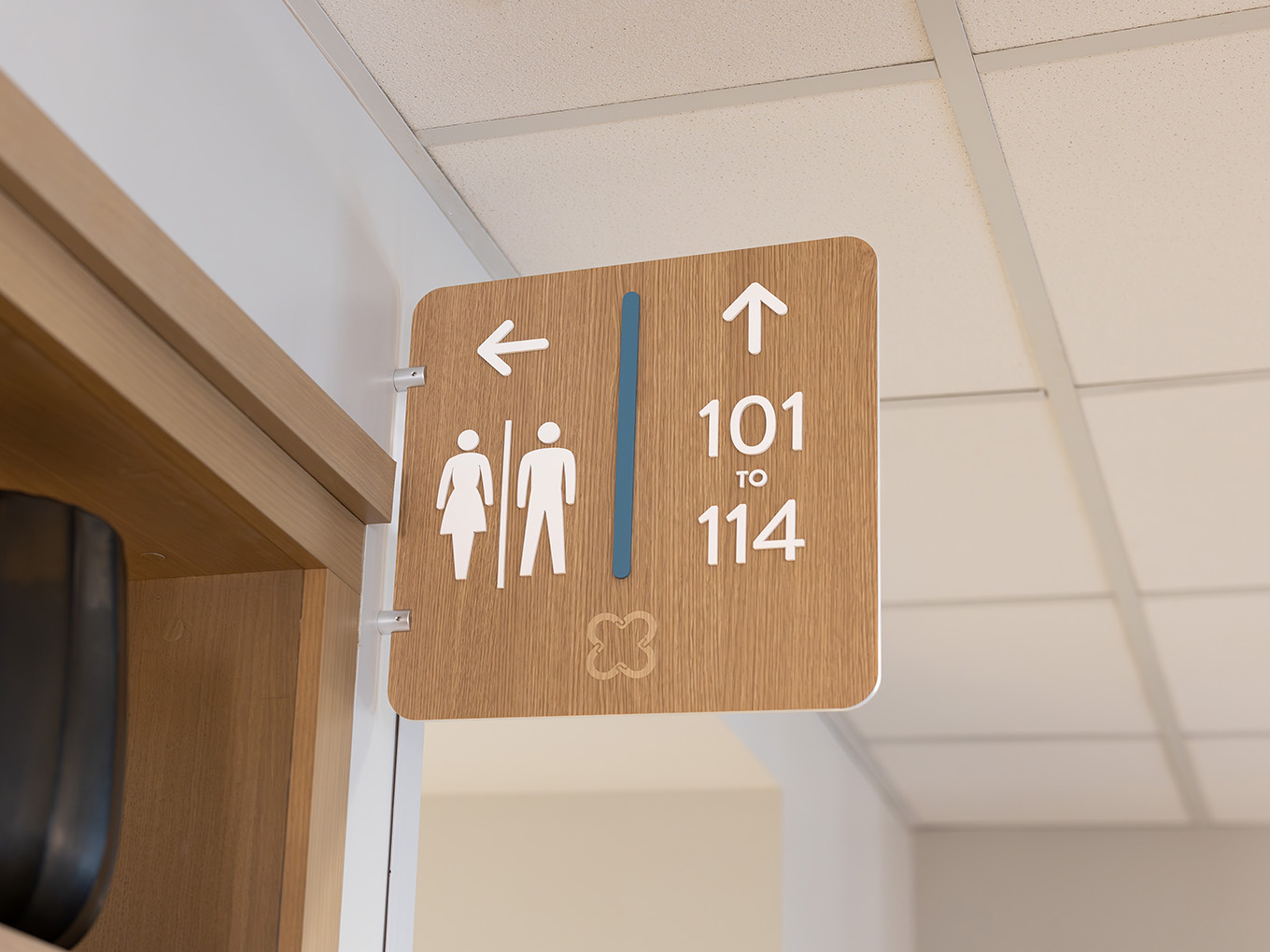 Wood-look wall-mounted directional sign at United Active Living Garrison Green pointing toward suites 101–114 and the washrooms, featuring dimensional arrows, washroom icons, a blue accent bar, and the engraved United floral logo.